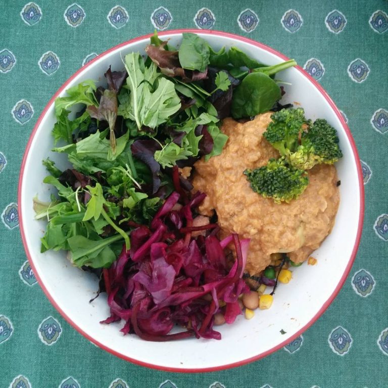 Bowl containing mixed salad leaves, red coleslaw and a thick peanut butter stew, with a couple of kernels of sweetcorn and peas