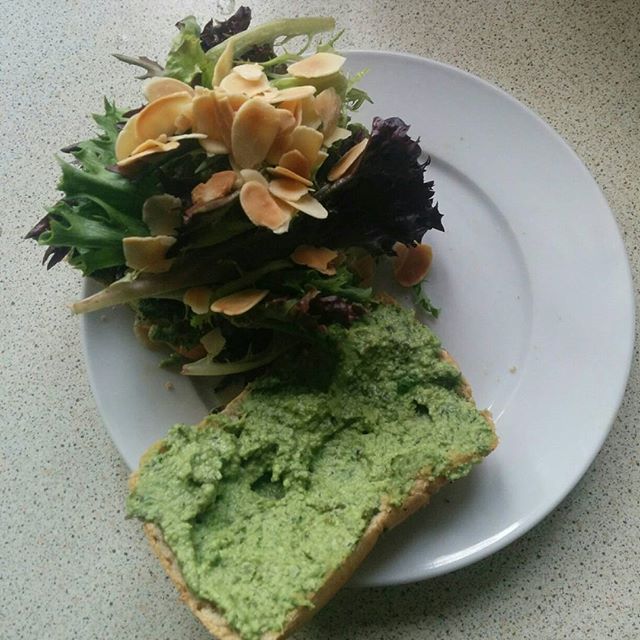 two slices of toast, one spread with a light green pesto spread, the other topped with mixed salad leaves and toasted flaked almonds