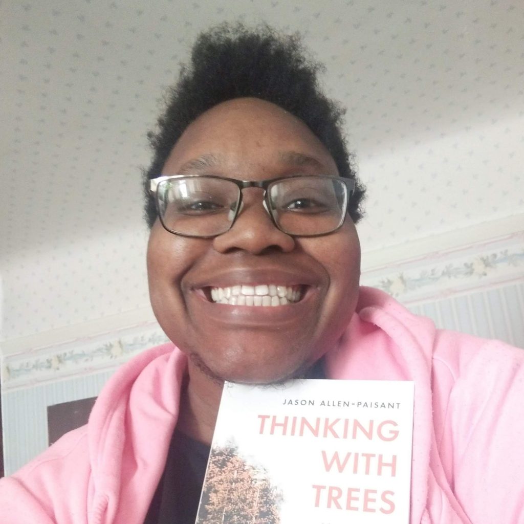 LiLi holding a copy of Thinking with Trees by Jason Allen-Paisant, smiling broadly and wearing a candy pink hoodie.